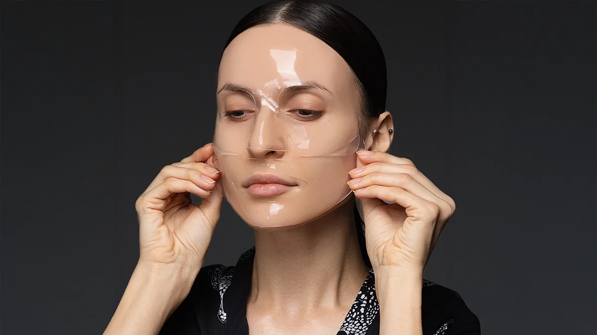 A beautiful young woman putting on FAQ Hydrogel Face Mask Glass Skin products on a dark background